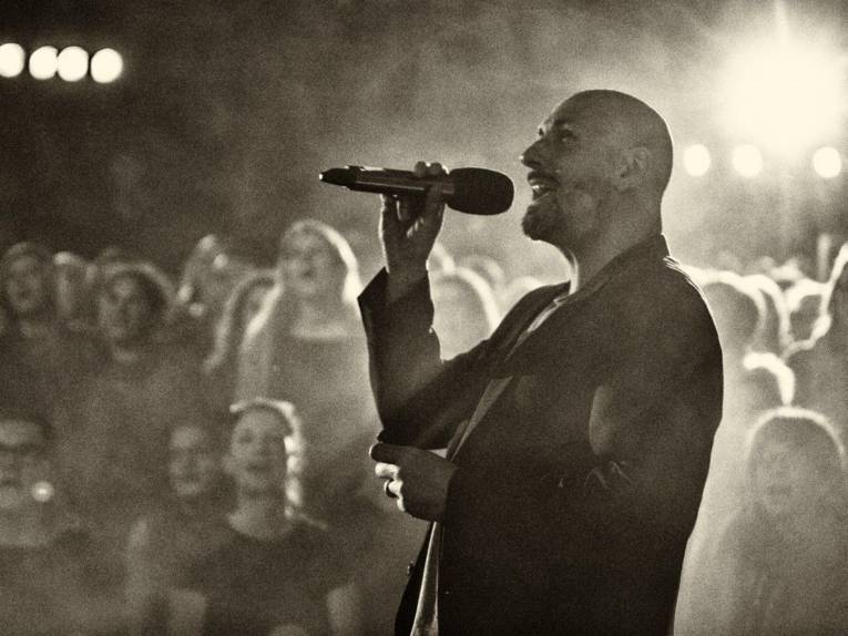 Mann singt in ein Mikrofon; im Hintergrund ist Publikum zu sehen, das mitsingt.